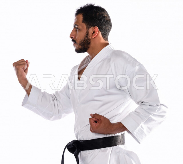 بورتريه بزاوية جانبية لرجل عربي خليجي سعودي يمارس رياضة الكاراتيه اليابانية، زي الكاراتيه الأبيض مع الحزام الأسود، التدرب على حركات قتالية خاصة، فنون قتالية يابانية، فنون الدفاع عن النفس، القوة البدنية، تطبيق حركات الكاتا، خلفية بيضاء