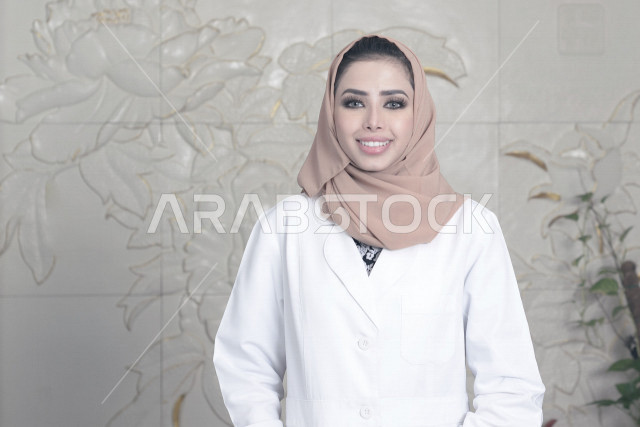 طبيبة عربية خليجية سعودية مبتسمة وسعيدة ترتدي المعطف الطبي وتنظر الى الكاميرا ، العمل في القطاع الصحي ،تقديم الخدمات الصحية، مفهوم الطب والرعاية الصحية