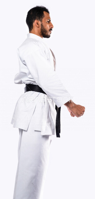 بورتريه بزاوية جانبية لرجل عربي خليجي سعودي يمارس رياضة الكاراتيه اليابانية، زي الكاراتيه الأبيض مع الحزام الأسود، التدرب على حركات قتالية خاصة، فنون قتالية يابانية، فنون الدفاع عن النفس، القوة البدنية، تطبيق حركات الكاتا، خلفية بيضاء