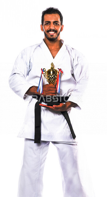 بورتريه لرجل عربي خليجي سعودي يرتدي زي الكاراتيه الأبيض مع الحزام الأسود ويمسك بيده كأس البطولة، ممارسة رياضة الكاراتيه اليابانية، تعبيرات وجه مبتسمة تدل على الثقة والقوة، الفوز بالمسابقة، خلفية بيضاء