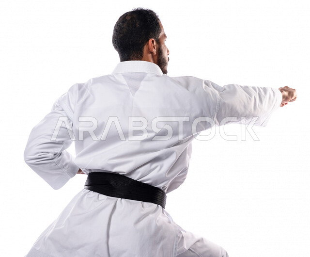 بورتريه من الخلف لرجل عربي خليجي سعودي يمارس رياضة الكاراتيه اليابانية، زي الكاراتيه الأبيض مع الحزام الأسود، التدرب على حركات قتالية خاصة، فنون قتالية يابانية، فنون الدفاع عن النفس، القوة البدنية، تطبيق حركات الكاتا، خلفية بيضاء