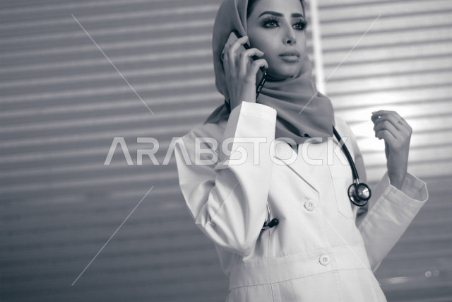 طبيبة عربية خليجية سعودية ترتدي المعطف الطبي و سماعة الطبيب ، تقوم باجراء مكالمة عبر الهاتف المحمول ، ايماءات وجه ويدين مختلفة ، تقديم الاستشارات الطبية والخدمات الصحية ، مفهوم الطب والرعاية الصحية