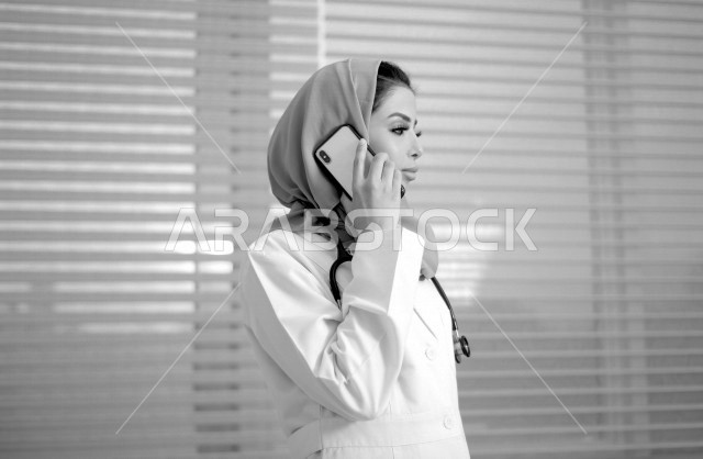 طبيبة عربية خليجية سعودية ترتدي المعطف الطبي و سماعة الطبيب ، تقوم باجراء مكالمة عبر الهاتف المحمول ، ايماءات وجه ويدين مختلفة ، تقديم الاستشارات الطبية والخدمات الصحية ، مفهوم الطب والرعاية الصحية