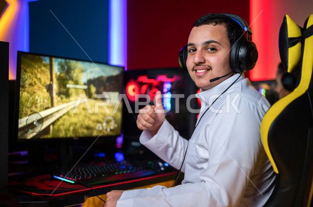 شاب عربي سعودي خليجي يرتدي سماعة الرأس و يلعب بألعاب الفيديو، إيماءات وجه تدل على الإندماج والتفاعل مع اللعبه، اللعب في مقهى ألعاب الكمبيوتر, المتعة و التسلية بألعاب الفيديو، ألعاب الكمبيوتر الترفيهية، تقنيات و ألعاب, أنشطه ترفيهيه