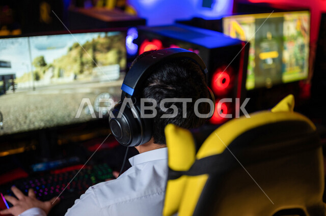  منظر من الخلف لشاب عربي سعودي خليجي يرتدي سماعة الرأس و يلعب بألعاب الفيديو، اللعب في مقهى ألعاب الكمبيوتر, المتعة و التسلية بألعاب الفيديو، ألعاب الكمبيوتر الترفيهية، تقنيات و ألعاب, أنشطه ترفيهيه