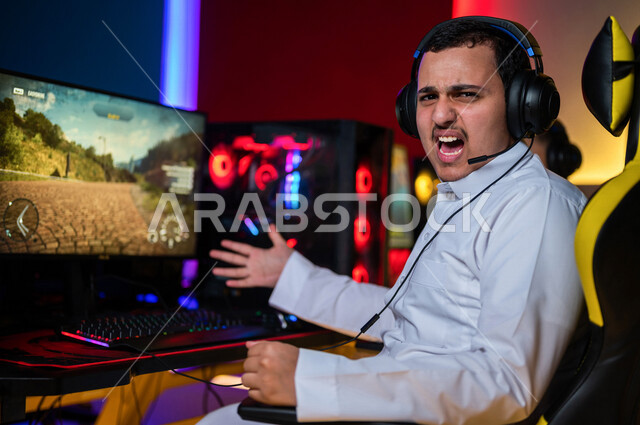 شاب عربي سعودي خليجي يرتدي سماعة الرأس و يلعب بألعاب الفيديو، إيماءات وجه تدل على الإندماج والتفاعل مع اللعبه، اللعب في مقهى ألعاب الكمبيوتر, المتعة و التسلية بألعاب الفيديو، ألعاب الكمبيوتر الترفيهية، تقنيات و ألعاب, أنشطه ترفيهيه