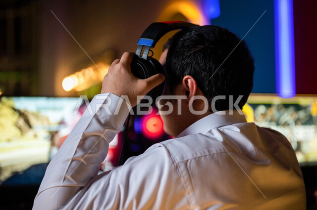  منظر من الخلف لشاب عربي سعودي خليجي يرتدي سماعة الرأس و يلعب بألعاب الفيديو، اللعب في مقهى ألعاب الكمبيوتر, المتعة و التسلية بألعاب الفيديو، ألعاب الكمبيوتر الترفيهية، تقنيات و ألعاب, أنشطه ترفيهيه