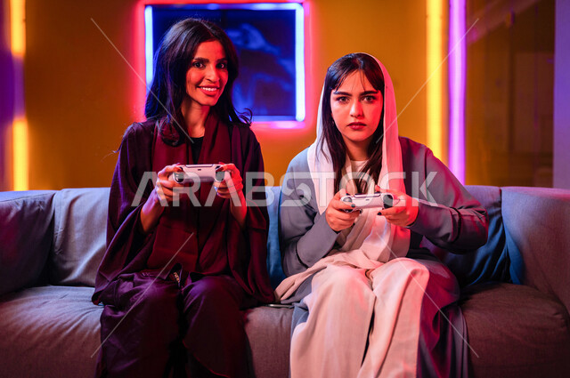 إمرأتان عربيتان سعوديتان خليجيتان تمسكان عصا التحكم و تلعبان بألعاب الفيديو، إيماءات وجه تدل على الإندماج والتفاعل مع اللعبه، المتعة و التسلية بألعاب الفيديو، ألعاب البلايستيشن الترفيهية، اللعب مع الأصدقاء، اللعب الجماعي, تسليه وترفيه, تقنيات و ألعاب