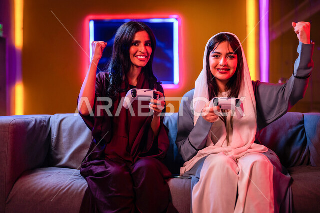 إمرأتان عربيتان سعوديتان خليجيتان تمسكان عصا التحكم و تلعبان بألعاب الفيديو، إيماءات وجه تدل على الإندماج والتفاعل مع اللعبه، المتعة و التسلية بألعاب الفيديو، ألعاب البلايستيشن الترفيهية، اللعب مع الأصدقاء، اللعب الجماعي, تسليه وترفيه, تقنيات و ألعاب