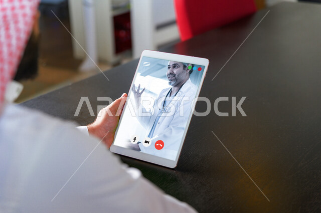 رجل عربي خليجي سعودي يحمل بيده جهاز التابليت، اجراءة مكالمة فيديو، التواصل مع الطبيب من خلال التابليت، القاء التحية من خلال شاشة الهاتف، تطبيق استشارات طبية، تقنيات حديثة، دردشة وتواصل، خدمات طبية، حجز موعد مع الطبيب عن بعد