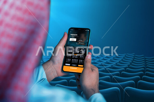 رجل عربي خليجي سعودي يجلس ويمسك بيده الهاتف المحمول، تصفح التطبيقات الالكترونية، تطبيق فينوم، الواجهة الامامية للتطبيقات الالكترونية، استخدام تقنيات حديثة، تطبيق يعرض مواعيد عرض افلام السينما، خدمات السينما، العروض التلفيزونية