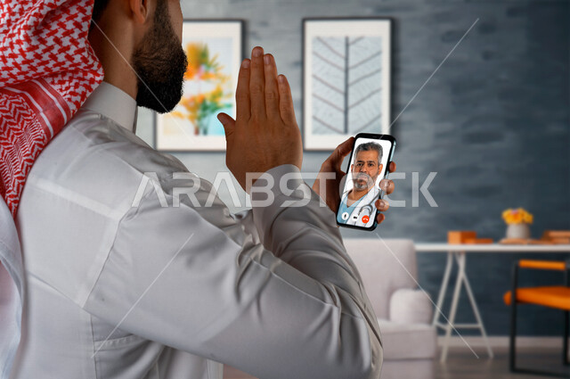 رجل عربي خليجي سعودي يحمل بيده الهاتف المحمول، اجراءة مكالمة فيديو، التواصل مع الطبيب من خلال الهاتف المحمول، القاء التحية من خلال شاشة الهاتف، تطبيق استشارات طبية، خدمات طبية، حجز موعد مع الطبيب عن بعد