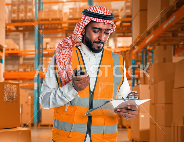 مهندس عربي خليجي سعودي يرتدي الزي السعودي التقليدي و سترة الحماية خاصة بالعمل ، يستخدم جهاز اتصال لاسلكي بإيماءات وجه تدل على التركيز ، يحمل بيده تقارير ، الإشراف على المشاريع ، إعطاء أوامر خاصة بالعمل ، مهن و وظائف هندسية ، ادارة المشاريع الهندسية 