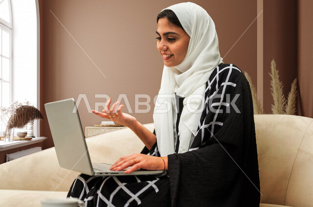 A smiling Saudi Arabian Gulf woman sitting in a room using a laptop ...