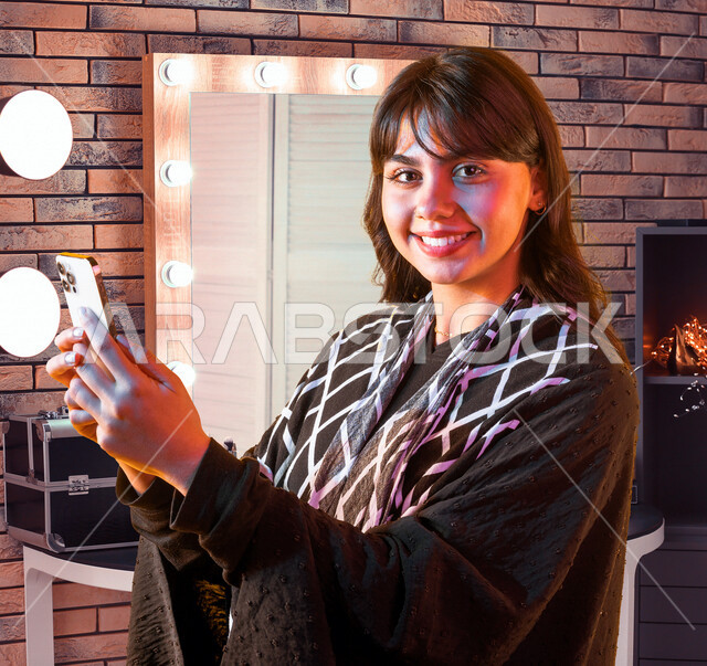 شابة عربية خليجية سعودية مبتسمة تحمل بيدها الهاتف المحمول تقوم بتصفح الإنترنت ، التسويق الإلكتروني ، إستخدام وسائل التواصل الإجتماعي ، إستخدام التطبيقات الذكية ، إجراء مكالمات فيديو و هاتفية ، أخذ صور تذكارية ، إستخدام أجهزة تقنية حديثة