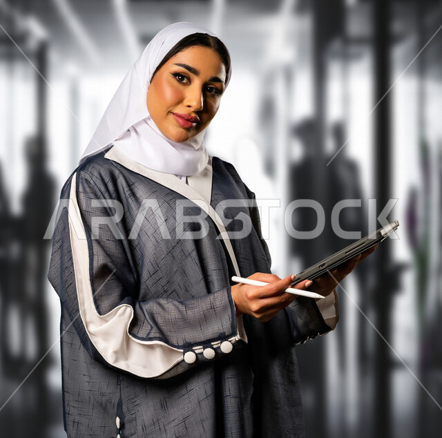 A smiling Saudi Gulf Arab businesswoman holding a technical device, a tablet and a pen, standing in a company specializing in following up business, a businesswoman, taking notes and reviewing them, supervising business, using technical devices at work