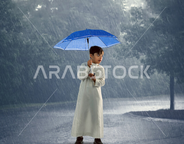 طفل عربي خليجي سعودي مصاب بمتلازمة داون، يحمل بيده مظلة باللون الأزرق يقف تحت المطر ، مظلة حامية من المطر، أصحاب الهمم، ذوي الاحتياجات الخاصة، خلفية للطبيعة الماطرة ، الشمسية