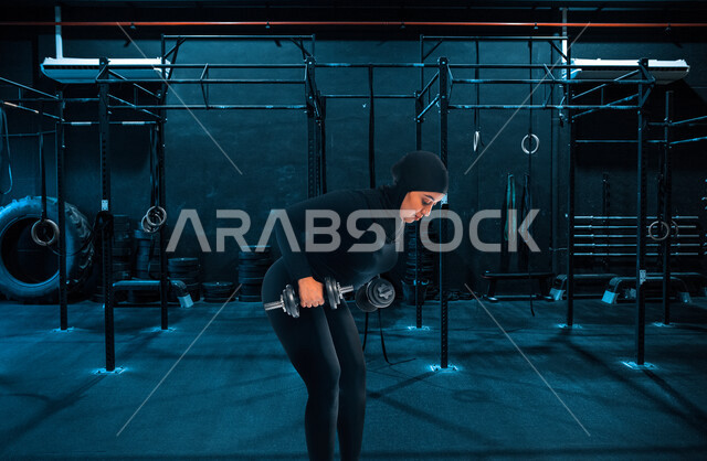 فتاة عربية سعودية محجبة في النادي الرياضي بالملابس الرياضية، ممارسة التمارين الرياضية مع الدمبل، نشاط بدني للمحافظة على الصحة واللياقة الجسدية، كمال الأجسام، أسلوب حياة صحي، بناء عضل، تمارين رفع الأثقال (دمبلز)، رفع الأوزان الحديدية الثقيلة
