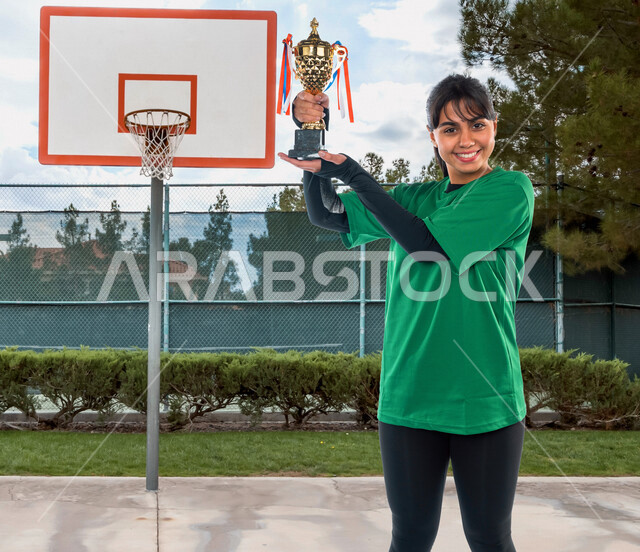 فتاة عربية خليجية سعودية، ترتدي الملابس الرياضية، تحمل بيدها كأس الفوز، بإيماءات تدل على السعادة بالفوز والنصر، ممارسة رياضة كرة السلة، رياضات الألعاب، أنشطة رياضية نسائية في السعودية، الفريق السعودي النسائي، المنتخب الوطني السعودي لكرة السلة
