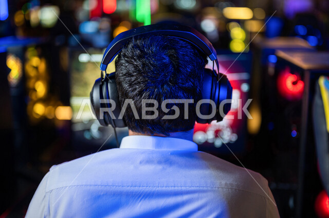  منظر من الخلف لشاب عربي سعودي خليجي يرتدي سماعة الرأس و يلعب بألعاب الفيديو، اللعب في مقهى ألعاب الكمبيوتر, المتعة و التسلية بألعاب الفيديو، ألعاب الكمبيوتر الترفيهية، تقنيات و ألعاب, أنشطه ترفيهيه