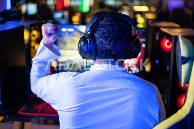  منظر من الخلف لشاب عربي سعودي خليجي يرتدي سماعة الرأس و يلعب بألعاب الفيديو، اللعب في مقهى ألعاب الكمبيوتر, المتعة و التسلية بألعاب الفيديو، ألعاب الكمبيوتر الترفيهية، تقنيات و ألعاب, أنشطه ترفيهيه