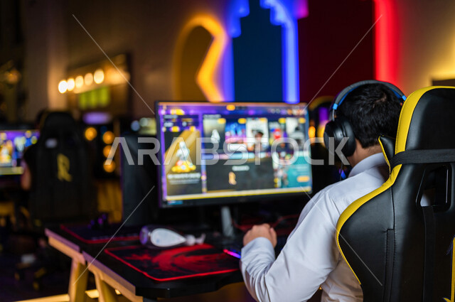  منظر من الخلف لشاب عربي سعودي خليجي يرتدي سماعة الرأس و يلعب بألعاب الفيديو، اللعب في مقهى ألعاب الكمبيوتر, المتعة و التسلية بألعاب الفيديو، ألعاب الكمبيوتر الترفيهية، تقنيات و ألعاب, أنشطه ترفيهيه