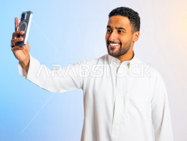 بورتريه لشاب عربي خليجي سعودي مبتسم وسعيد يستخدم الهاتف المحمول ، بايماءات تد على اجراء مكالمة فيديو ، بث مباشر ، التقاط الصور التذكارية ، تصوير مقاطع فيديو ،  التواصل الاجتماعي