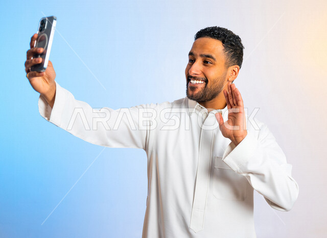بورتريه لشاب عربي خليجي سعودي مبتسم وسعيد يستخدم الهاتف المحمول ، بايماءات تد على اجراء مكالمة فيديو ، بث مباشر ، التقاط الصور التذكارية ، تصوير مقاطع فيديو ،  التواصل الاجتماعي