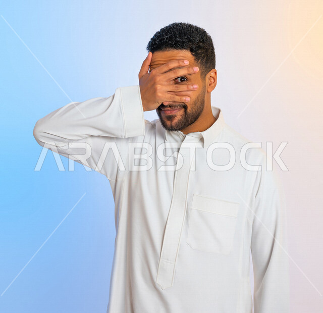 Portrait of a Saudi Arabian Gulf man with hand gestures indicating hiding and peeking, a shy man hiding his face with his hand and looking with one eye, peeking, observing something