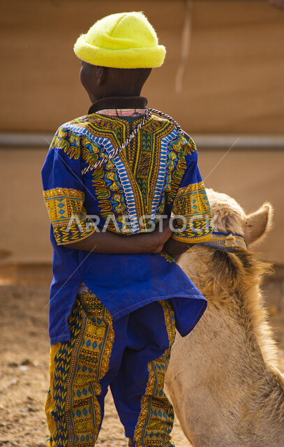 صورة من الخلف لطفل يقف بجانب حيوان الجمل في المملكة العربية السعودية، تربية الابل، حيوان الجمل، طفل اسمر