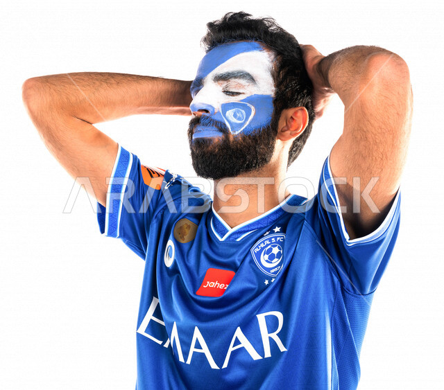 بورتريه لشاب عربي خليجي سعودي يرتدي تيشرت نادي الهلال السعودي ويضع يده على رأسه، تشجيع فريق الهلال السعودي لكرة قدم، متابعة مباراة كرة قدم، شاب يرسم على وجهه شعار شعار نادي الهلال السعودي، إيماءات وجه ويدين تدل على الدهشة والذهول والخذلان، خلفية بيضاء