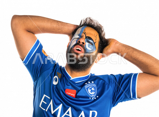 بورتريه لشاب عربي خليجي سعودي يرتدي تيشرت نادي الهلال السعودي ويضع يده على رأسه، تشجيع فريق الهلال السعودي لكرة قدم، متابعة مباراة كرة قدم، شاب يرسم على وجهه شعار شعار نادي الهلال السعودي، إيماءات وجه ويدين تدل على الدهشة والذهول والخذلان، خلفية بيضاء