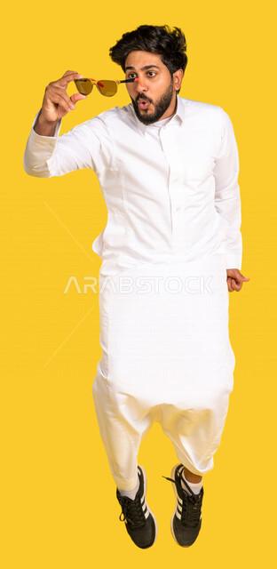 رجل عربي سعودي خليجي يقفز فرحا, ارتداء النظاره الشمسيه, اختيار نظاره جديده, اقتناء نظارات حماية من الشمس, صورة للجسم بالطول الكامل, خلفيه صفراء