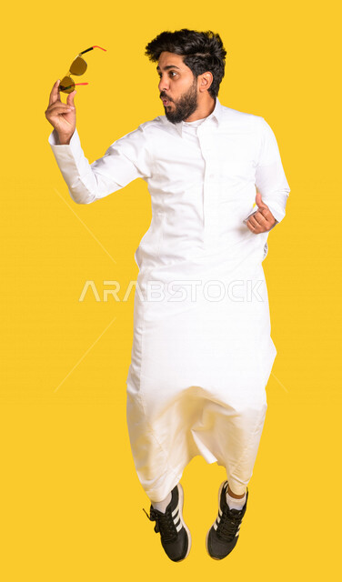 رجل عربي سعودي خليجي يقفز فرحا, ارتداء النظاره الشمسيه, اختيار نظاره جديده, اقتناء نظارات حماية من الشمس, صورة للجسم بالطول الكامل, خلفيه صفراء