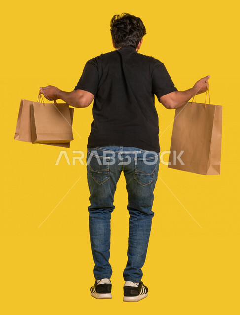 بورتريه من الخلف لرجل عربي خليجي سعودي يقفز في الهواء ويحمل بيديه أكياس التسوق ، الاستمتاع بالشراء والتسوق ، التسوق اونلاين ، خدمة التوصيل للمنزل ، استلام الطرود، خدمة الشحن والتوصيل ، صورة بالطول الكامل ، كامل الجسم 