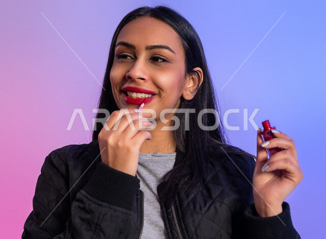 بورتريه لمرأة عربية خليجية سعودية تحمل إحدى أدوات التجميل (قلم أحمر الشفاة) ، خلفية ملونة لمرأة خبيرة بمستحضرات التجميل، التسويق لمنتجات التجميل، متجر بيع مستحضرات التجميل عبر الإنترنت ، مفهوم الإهتمام بالجمال والبشرة، ادوات مكياج تتوريال