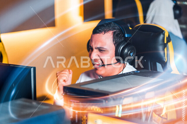 شاب عربي سعودي خليجي يرتدي سماعة الرأس و يلعب بألعاب الفيديو، إيماءات وجه تدل على الفوز، اللعب في مقهى ألعاب الكمبيوتر, المتعة و التسلية بألعاب الفيديو، ألعاب الكمبيوتر الترفيهية، تقنيات و ألعاب, أنشطه ترفيهيه, خلفيات هولوجراميه ثلاثية الأبعاد