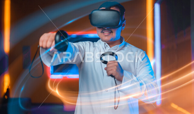 شاب عربي سعودي خليجي يرتدي نظارة العالم الافتراضي, يمسك بيده عصا التحكم و يلعب بألعاب العالم الإفتراضي، المتعة و التسلية بألعاب العالم الإفتراضي, تقنيات و ألعاب, قضاء وقت ممتع, أنشطه ترفيهيه, خلفيات هولوجراميه ثلاثية الأبعاد