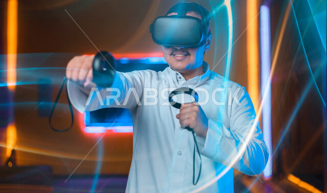 شاب عربي سعودي خليجي يرتدي نظارة العالم الافتراضي, يمسك بيده عصا التحكم و يلعب بألعاب العالم الإفتراضي، المتعة و التسلية بألعاب العالم الإفتراضي, تقنيات و ألعاب, قضاء وقت ممتع, أنشطه ترفيهيه, خلفيات هولوجراميه ثلاثية الأبعاد