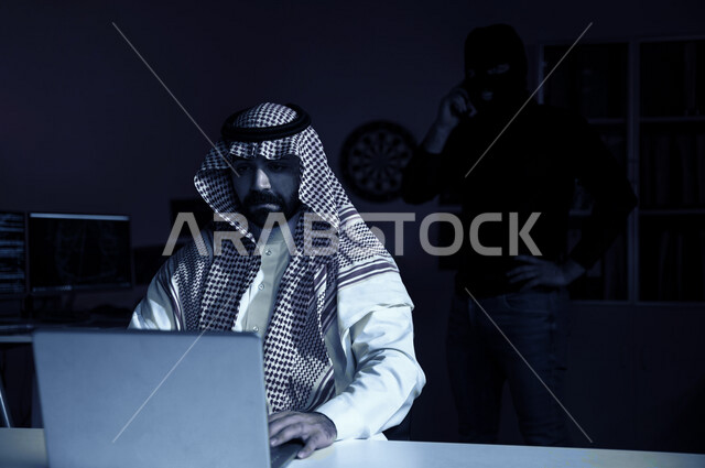 رجل أعمال عربي خليجي سعودي يجلس امام الحاسوب المحمول وخلفه رجل هكر ، العمل في مجال الامن السيبراني، الحماية من الهجمات الالكترونية ، الحفاظ على البيانات الشخصية و الدخول الآمن ،حماية المعلومات 