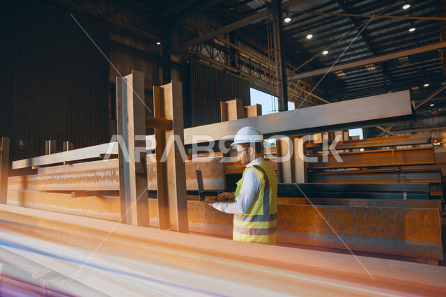 A Saudi Arabian Gulf engineer inside the iron factory, checking the quality of iron through his laptop, using engineering applications and programs, wearing a work helmet and protective jacket, the concept of engineering and industry
