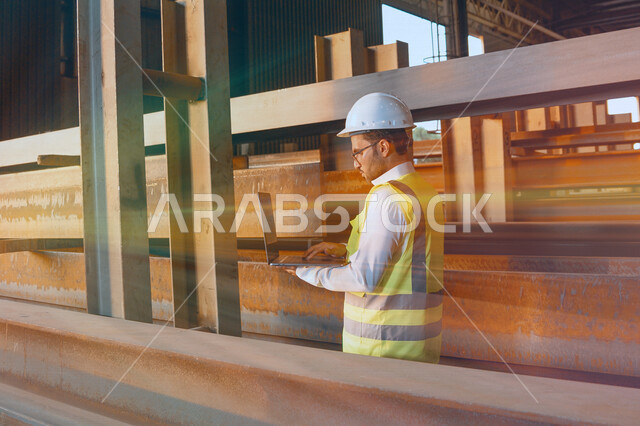 مهندس عربي خليجي سعودي داخل مصنع الحديد ، يفحص جودة الحديد من خلال الكمبيوتر المحمول ، استخدام التطبيقات والبرامج الهندسية ، يرتدي خوذة و سترة الحماية الخاصة بالعمل ، مفهوم الهندسة و الصناعة