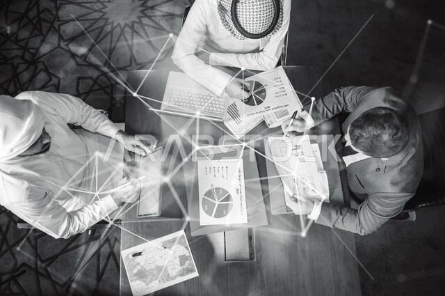 Black and white photo of a group of Saudi Gulf employees in a meeting inside a Saudi company, using a laptop computer to follow the stock exchange, analyze sales data and an economic growth chart. Business and money strategy
