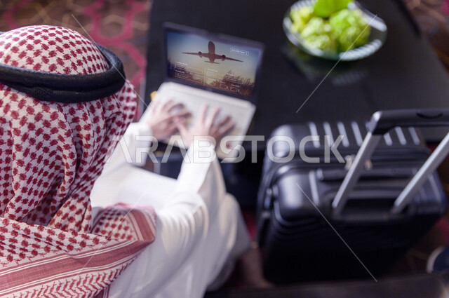 Photography from the back of a Saudi Arabian Gulf man using a laptop with his travel bag next to him, searching for travel offices and airlines, cutting travel tickets through the Internet, booking flights, booking flights