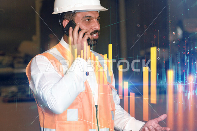مهندس عربي خليجي سعودي يرتدي خوذة الحماية وسترة الوقاية ، اجراء مكالمة من خلال الهاتف المحمول ، اتصال خاص بالعمل ، اعطاء الاوامر والتعليمات ، خلفية لمخطط بياني بتقنية الهولوجرام