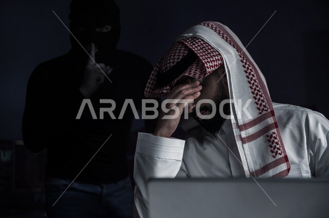 رجل عربي خليجي سعودي يجلس امام الحاسوب المحمول بايماءات تدل على الخسارة وخلفه رجل هكر ،التعرض لسرقة كلمات المرور والبيانات الشخصية ، اختراق النظام والبيانات، جرائم الكترونية ،الهجمات الالكترونية 