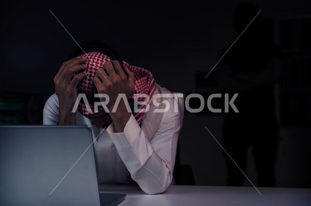 رجل عربي خليجي سعودي يجلس امام الحاسوب المحمول بايماءات تدل على الخسارة وخلفه رجل هكر ،التعرض لسرقة كلمات المرور والبيانات الشخصية ، اختراق النظام والبيانات، جرائم الكترونية ،الهجمات الالكترونية 