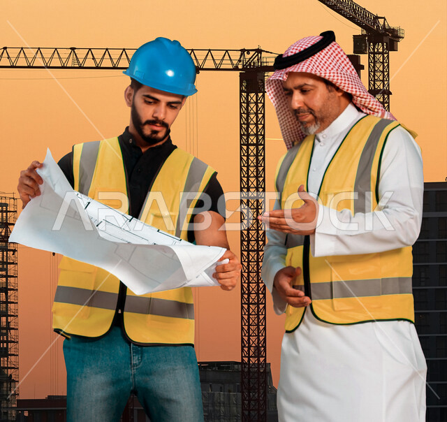 مهندسان معماريان سعوديان خليجيان يناقشان مخطط لمشروع البناء، إرتداء سترة الحماية الخاصة بالعمل, تدقيق العمل، الإشراف والنقاش لمشاريع قيد الانشاء، مفهوم الهندسة المعمارية وادارة المشاريع ، مهن ووظائف هندسية