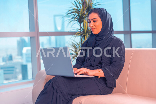 مرأة عربية خليجية سعودية، تقوم بإستخدام جهاز الحاسوب المحمول، العمل عن بعد، جلسة عمل، مهن ووظائف، إستخدام أجهزة تقنية حديثة، التسويق الإلكتروني، خلفية لأبراج الرياض، تصفح الإنترنت، إستخدام وسائل التواصل الإجتماعي