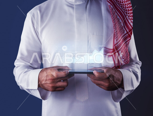 صورة مقربة ليد رجل عربي خليجي سعودي يقوم بإستخدام الهاتف المحمول، إستخدام تقنية الهولوجرام ثلاثية الأبعاد، الرسومات البيانية للبورصة المالية، متابعة سوق الاوراق المالية، الإستثمار والتداول المالي في البورصة، مراقبة الاقتصاد وسوق العملات الرقمية